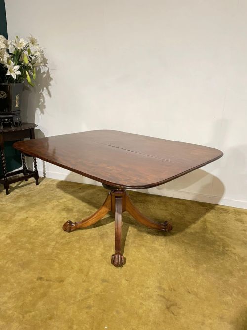 fig 3. A late Regency mahogany tilt-top breakfast table featuring a rectangular polished top with rounded corners, supported by a central pedestal and reeded quadruped legs with brass paw feet.
