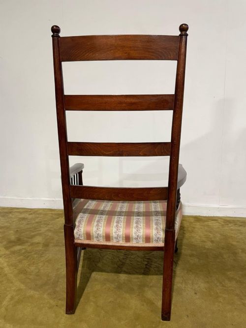 fig 5. An Arts and Crafts dark beech ladder back armchair featuring turned spindle arm supports, top finials, and a striped fabric upholstered seat.