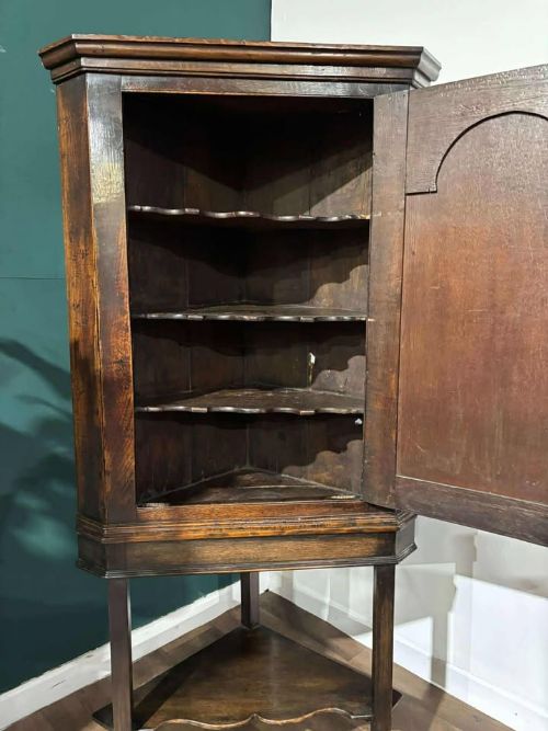 fig 3. An 18th-century dark oak corner cabinet featuring an arched fielded panel door, raised on a tall wooden stand with a lower shaped shelf.