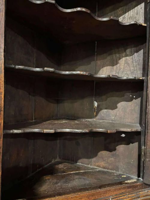 fig 8. An 18th-century dark oak corner cabinet featuring an arched fielded panel door, raised on a tall wooden stand with a lower shaped shelf.