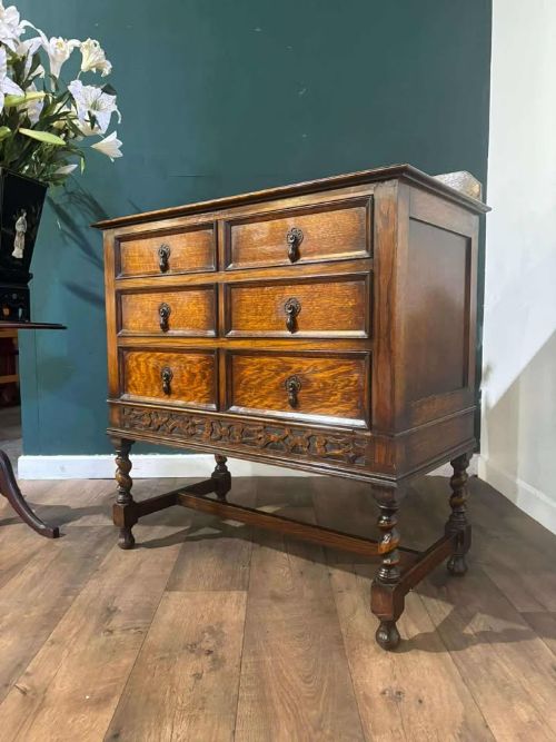 fig 6. A medium brown vintage oak chest of drawers featuring three split-panel drawer fronts, carved frieze detailing, a low back gallery, and barley twist legs connected by stretchers.