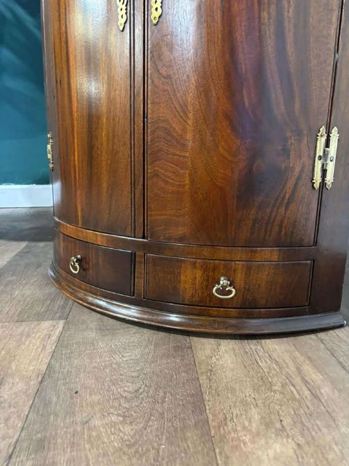 fig 1. A bow-fronted dark mahogany corner wall cupboard featuring twin doors with brass H-hinges and two drawers at the base.