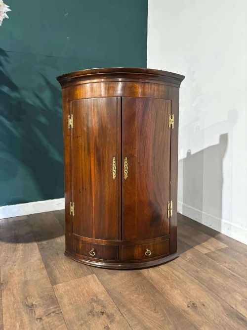 fig 2. A bow-fronted dark mahogany corner wall cupboard featuring twin doors with brass H-hinges and two drawers at the base.