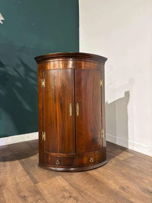 fig 3. A bow-fronted dark mahogany corner wall cupboard featuring twin doors with brass H-hinges and two drawers at the base.