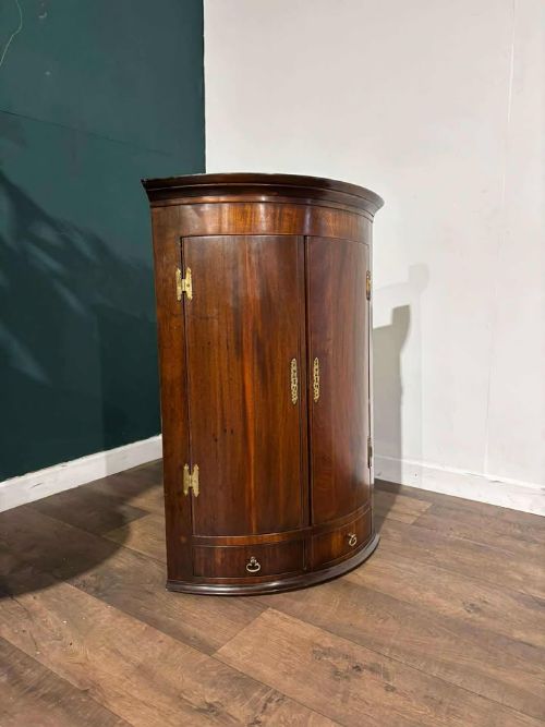 fig 4. A bow-fronted dark mahogany corner wall cupboard featuring twin doors with brass H-hinges and two drawers at the base.