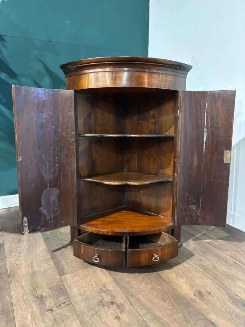 fig 7. A bow-fronted dark mahogany corner wall cupboard featuring twin doors with brass H-hinges and two drawers at the base.