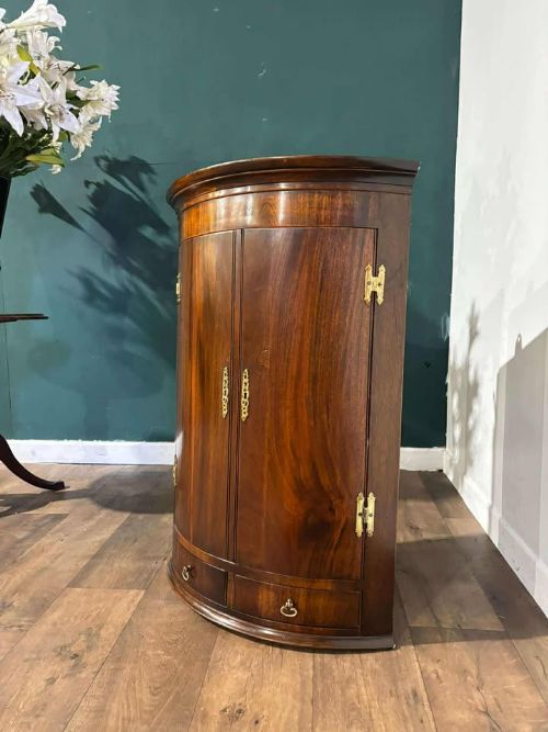 fig 8. A bow-fronted dark mahogany corner wall cupboard featuring twin doors with brass H-hinges and two drawers at the base.