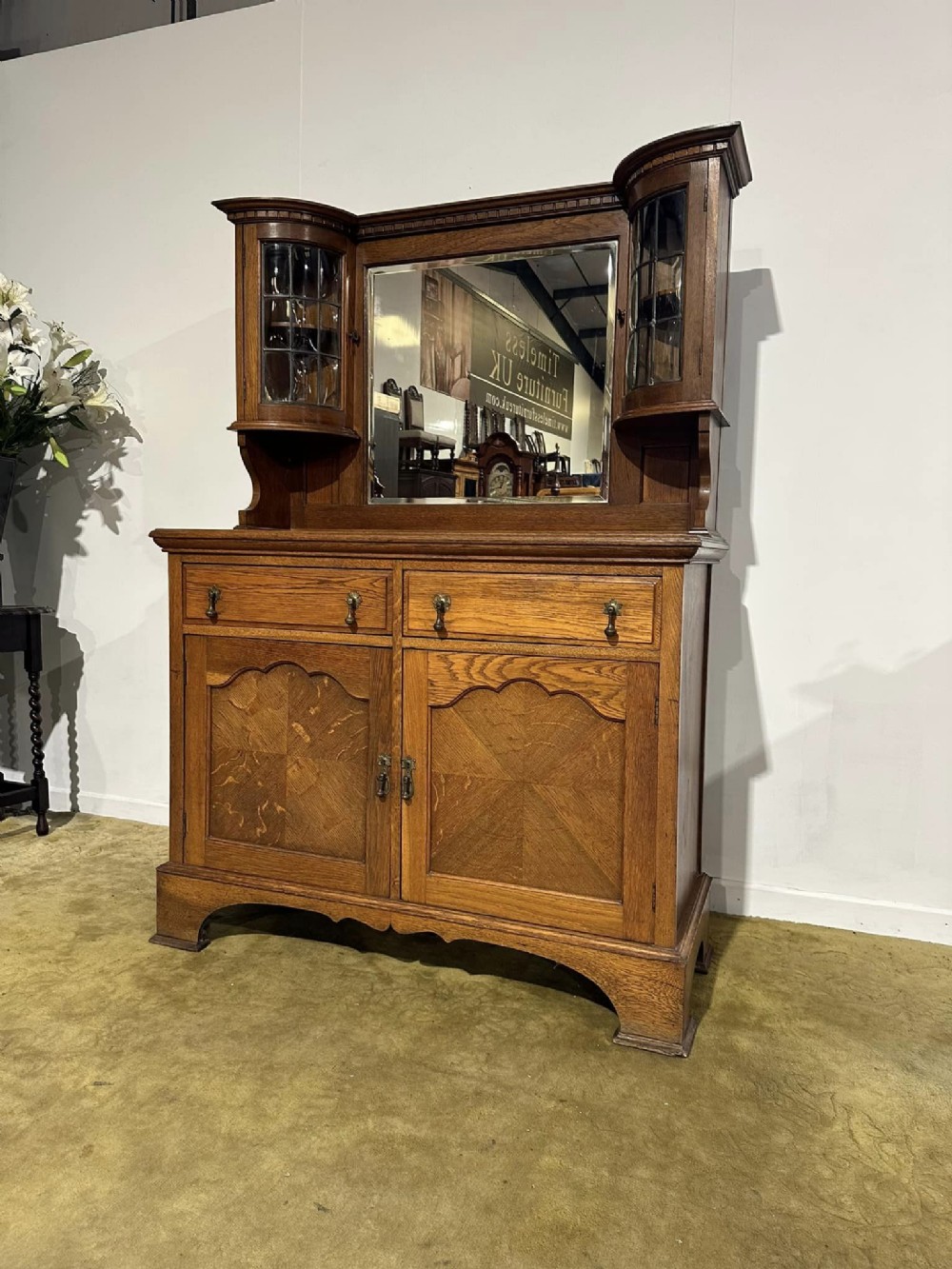A golden oak sideboard featuring a central mirror back flanked by two curved leaded glass cabinets above two drawers and two cupboard doors.