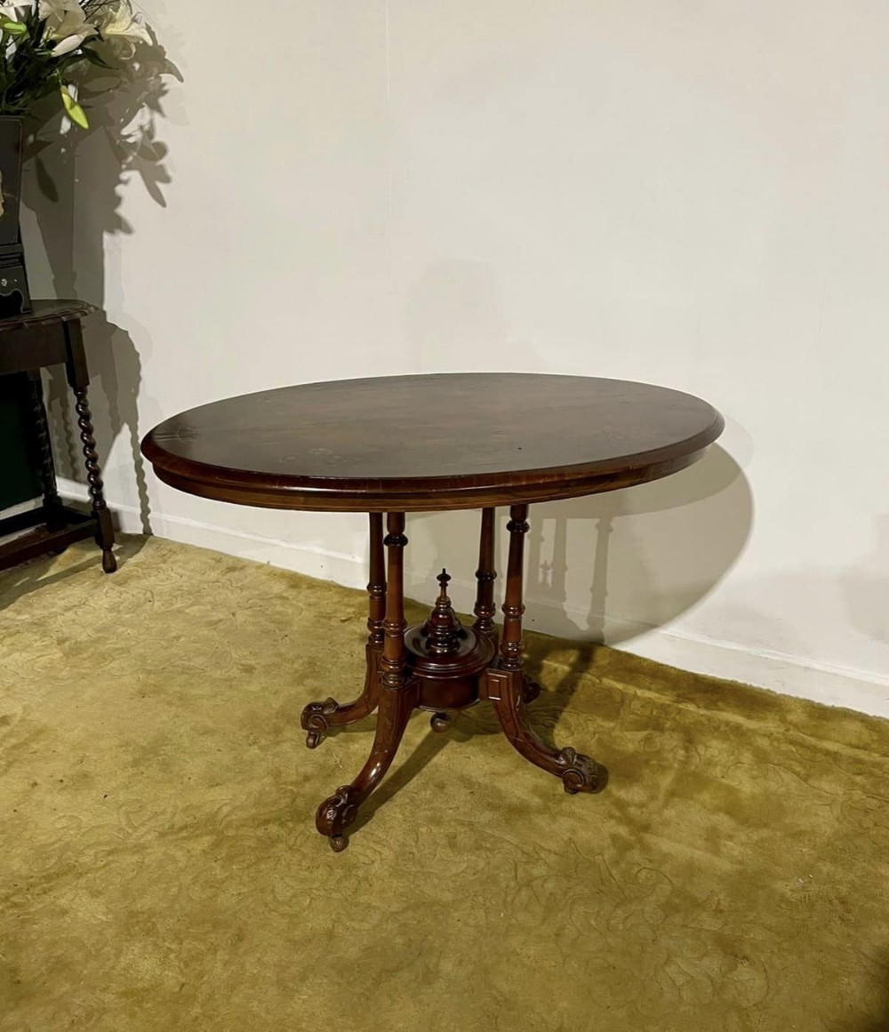 Victorian oval burr walnut occasional table featuring four turned column supports, a central finial, and carved legs on castors.