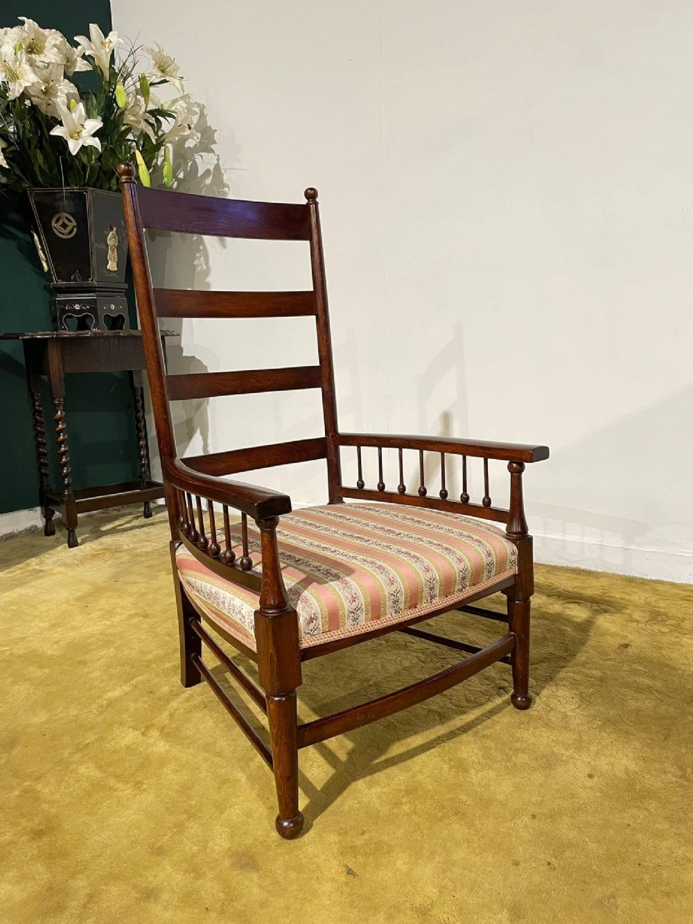 An Arts and Crafts dark beech ladder back armchair featuring turned spindle arm supports, top finials, and a striped fabric upholstered seat.