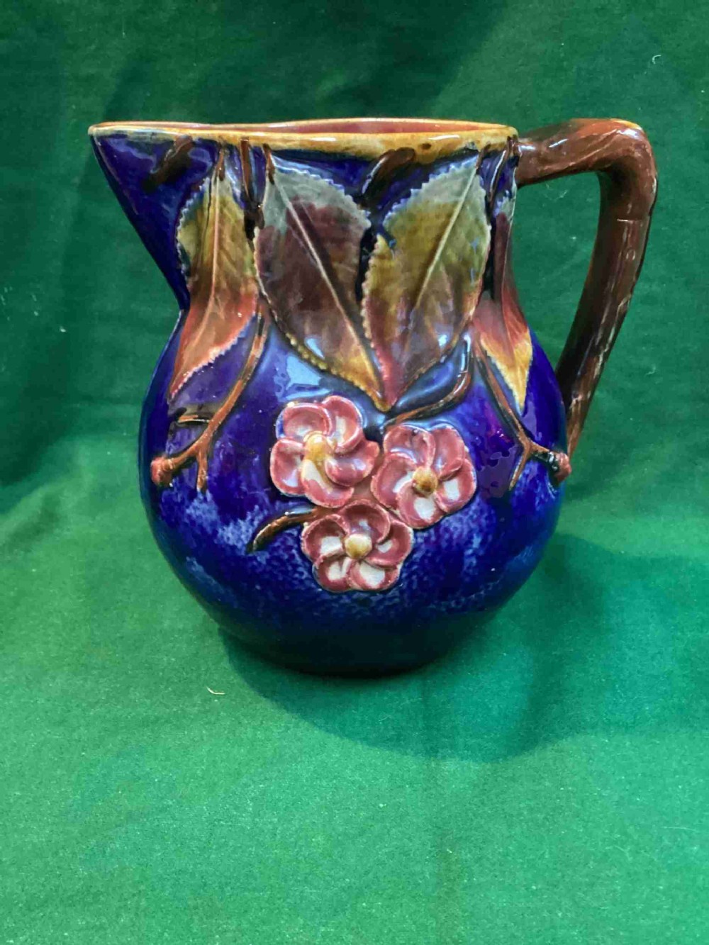 A Victorian majolica earthenware jug featuring a vibrant cobalt blue body adorned with relief-moulded green leaves, pink blossoms, and a faux-twining wood handle.