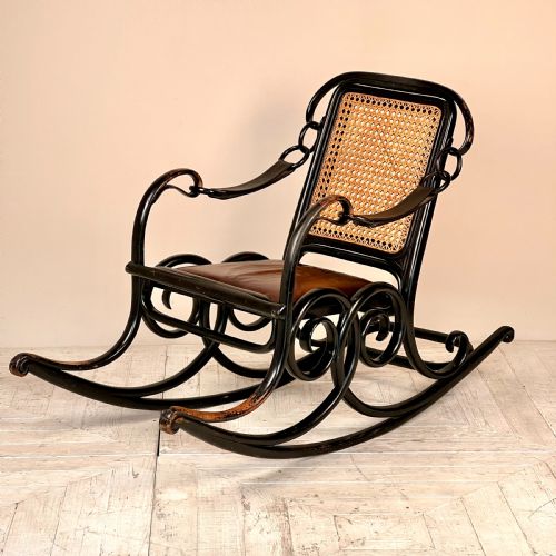 fig 1. A dark brown steam-bent beechwood rocking chair featuring elaborate side scrollwork, a caned backrest, leather arm straps, and a dark leather padded seat.