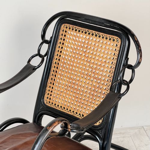 fig 2. A dark brown steam-bent beechwood rocking chair featuring elaborate side scrollwork, a caned backrest, leather arm straps, and a dark leather padded seat.
