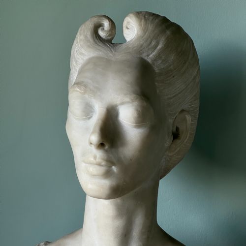 fig 1. A carved off-white marble bust depicting a lady with styled hair and a serene expression, mounted on a matching square marble plinth.