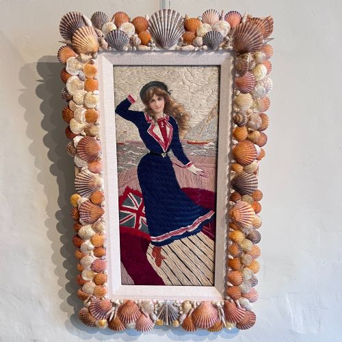 fig 1. A framed Victorian woolwork picture depicting a sailor girl standing on a ship deck beside a Union Jack flag, enclosed within a rectangular frame decorated with natural sea shells.