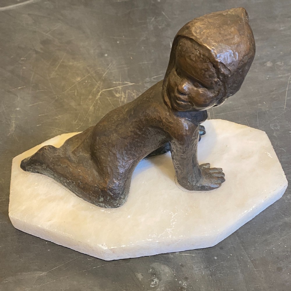 A small patinated bronze figurine depicting a crawling child wearing a bonnet, mounted on a hexagonal white alabaster base.