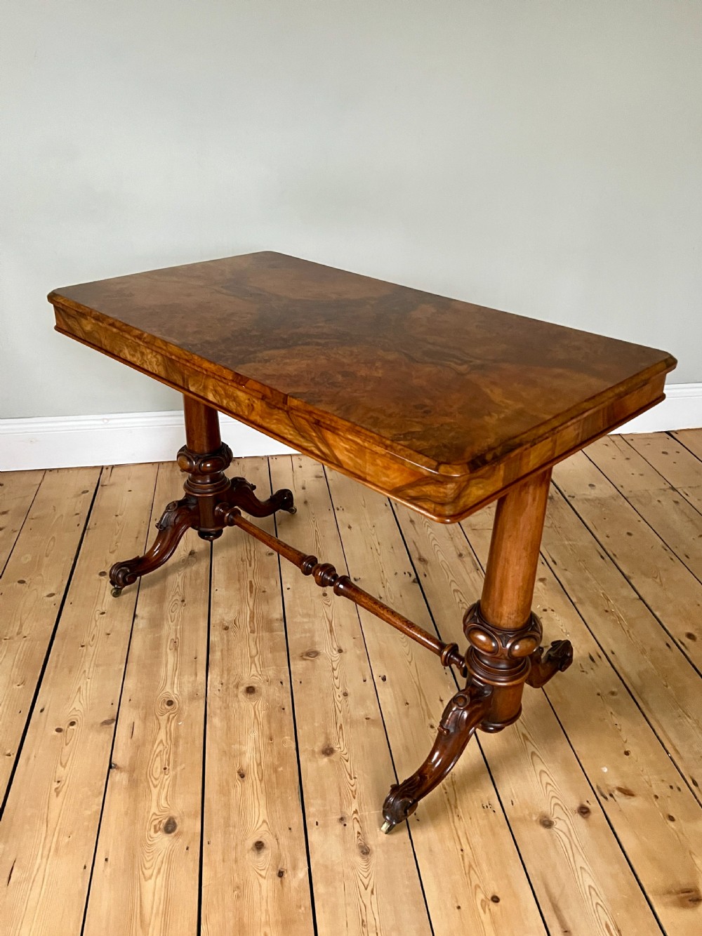 A Victorian burr walnut occasional table featuring a highly figured wood grain top, dual turned pedestals connected by a stretcher, and carved splayed legs on brass castors. View IV