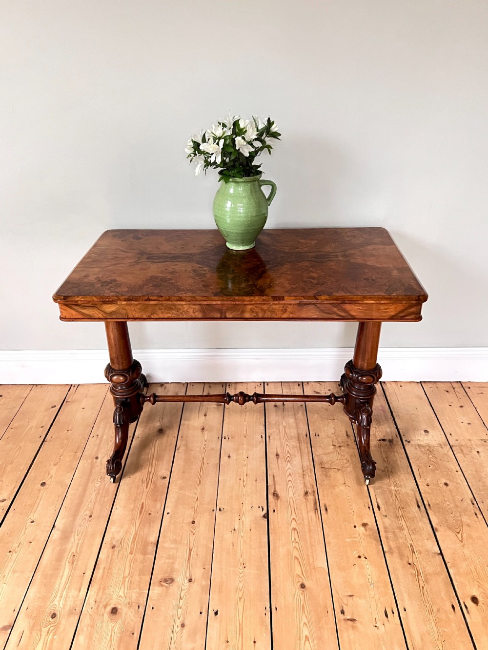 A Victorian burr walnut occasional table featuring a highly figured wood grain top, dual turned pedestals connected by a stretcher, and carved splayed legs on brass castors. View V