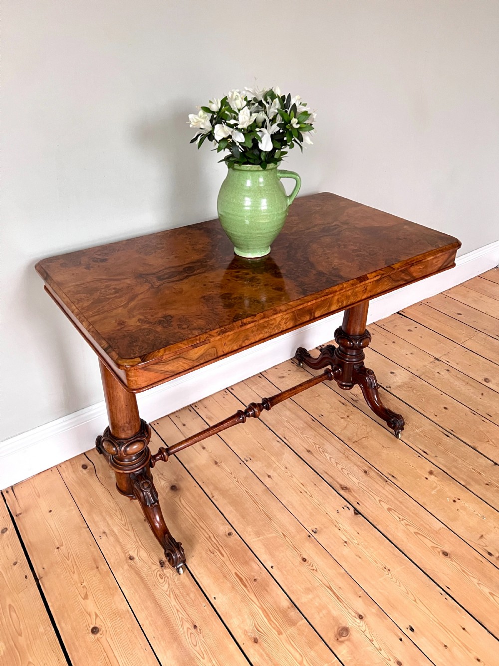 A Victorian burr walnut occasional table featuring a highly figured wood grain top, dual turned pedestals connected by a stretcher, and carved splayed legs on brass castors. View VI