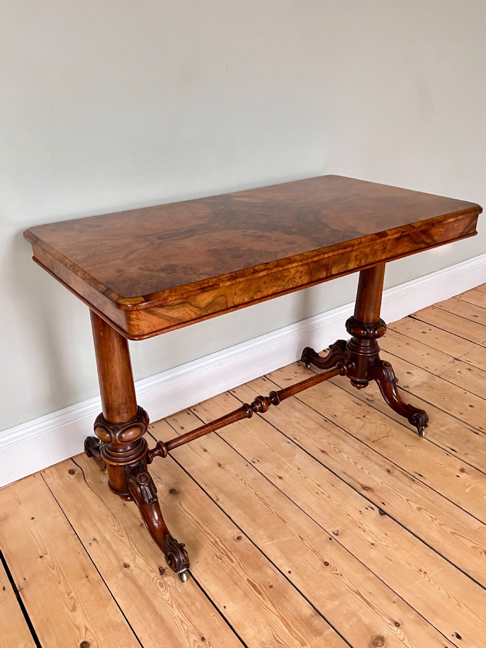 A Victorian burr walnut occasional table featuring a highly figured wood grain top, dual turned pedestals connected by a stretcher, and carved splayed legs on brass castors. View VII