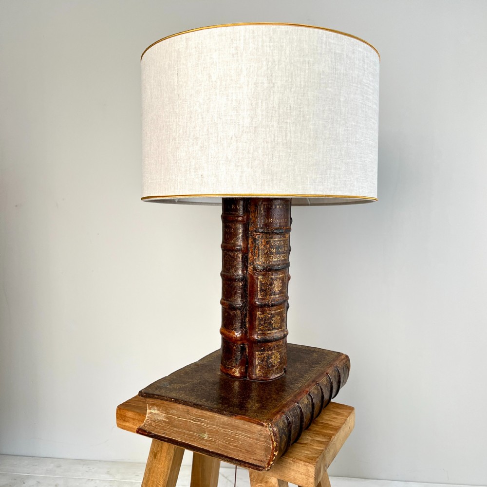 A table lamp featuring a column and base crafted from antique leather-bound book spines topped with a neutral drum shade.