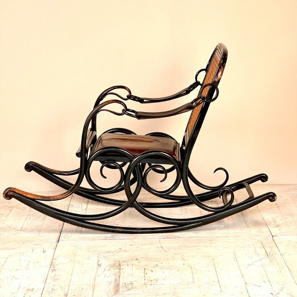 A dark brown steam-bent beechwood rocking chair featuring elaborate side scrollwork, a caned backrest, leather arm straps, and a dark leather padded seat.