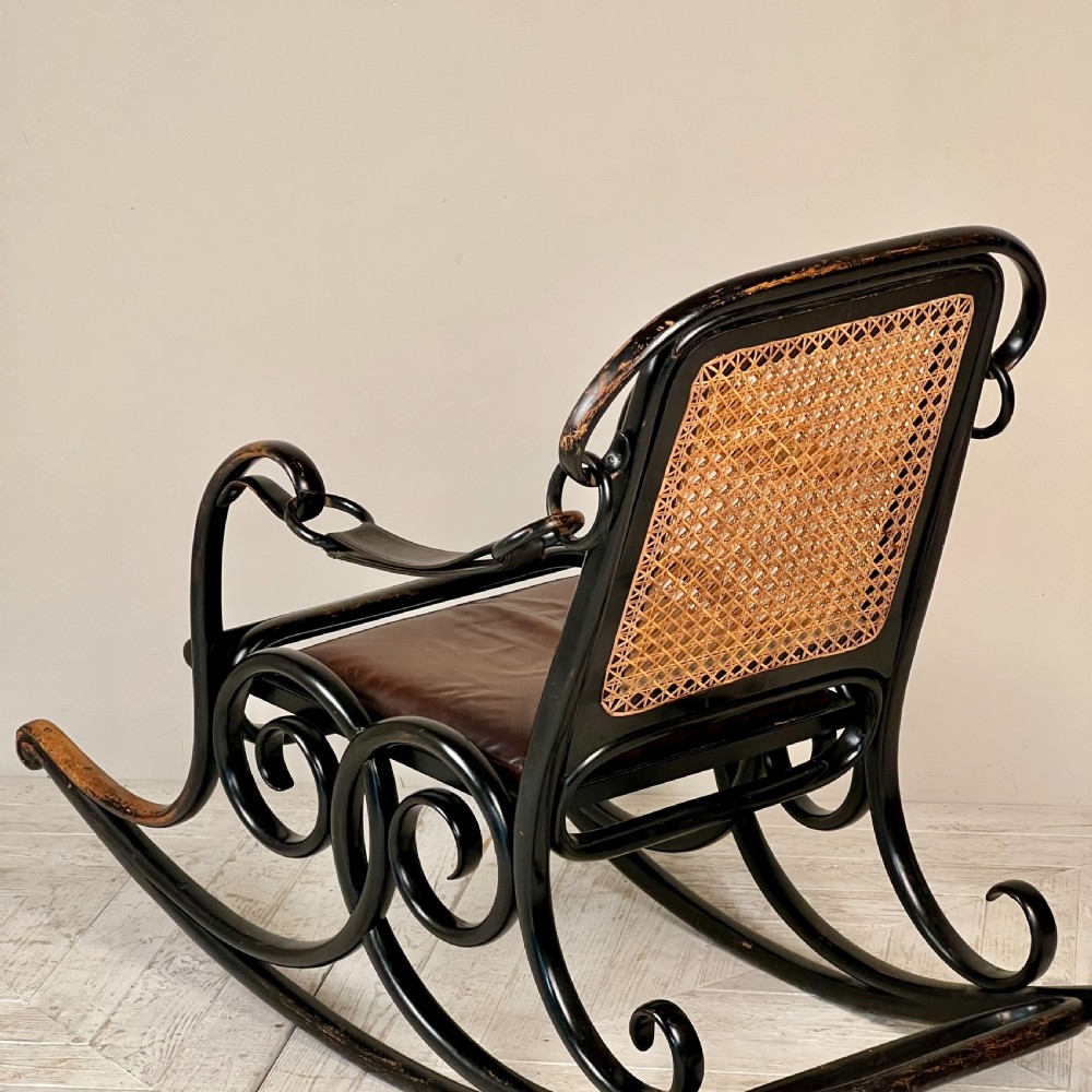 A dark brown steam-bent beechwood rocking chair featuring elaborate side scrollwork, a caned backrest, leather arm straps, and a dark leather padded seat. View IV