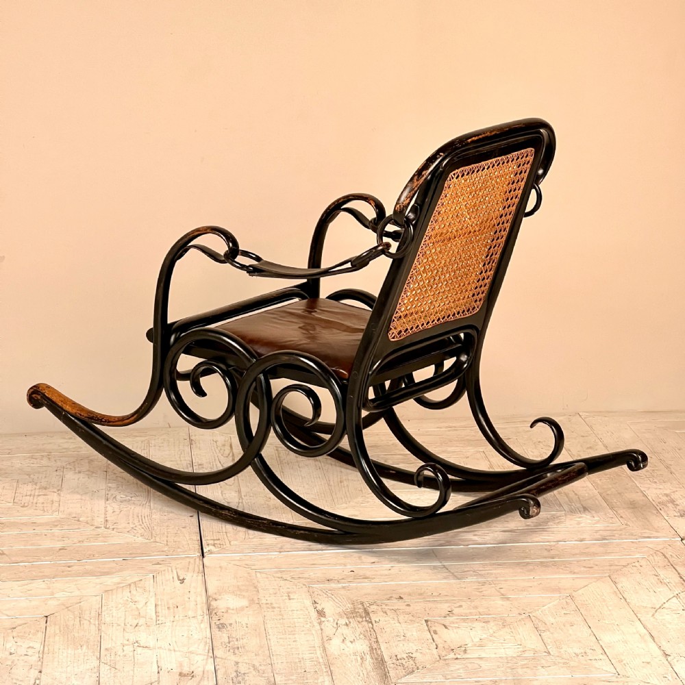 A dark brown steam-bent beechwood rocking chair featuring elaborate side scrollwork, a caned backrest, leather arm straps, and a dark leather padded seat. View V