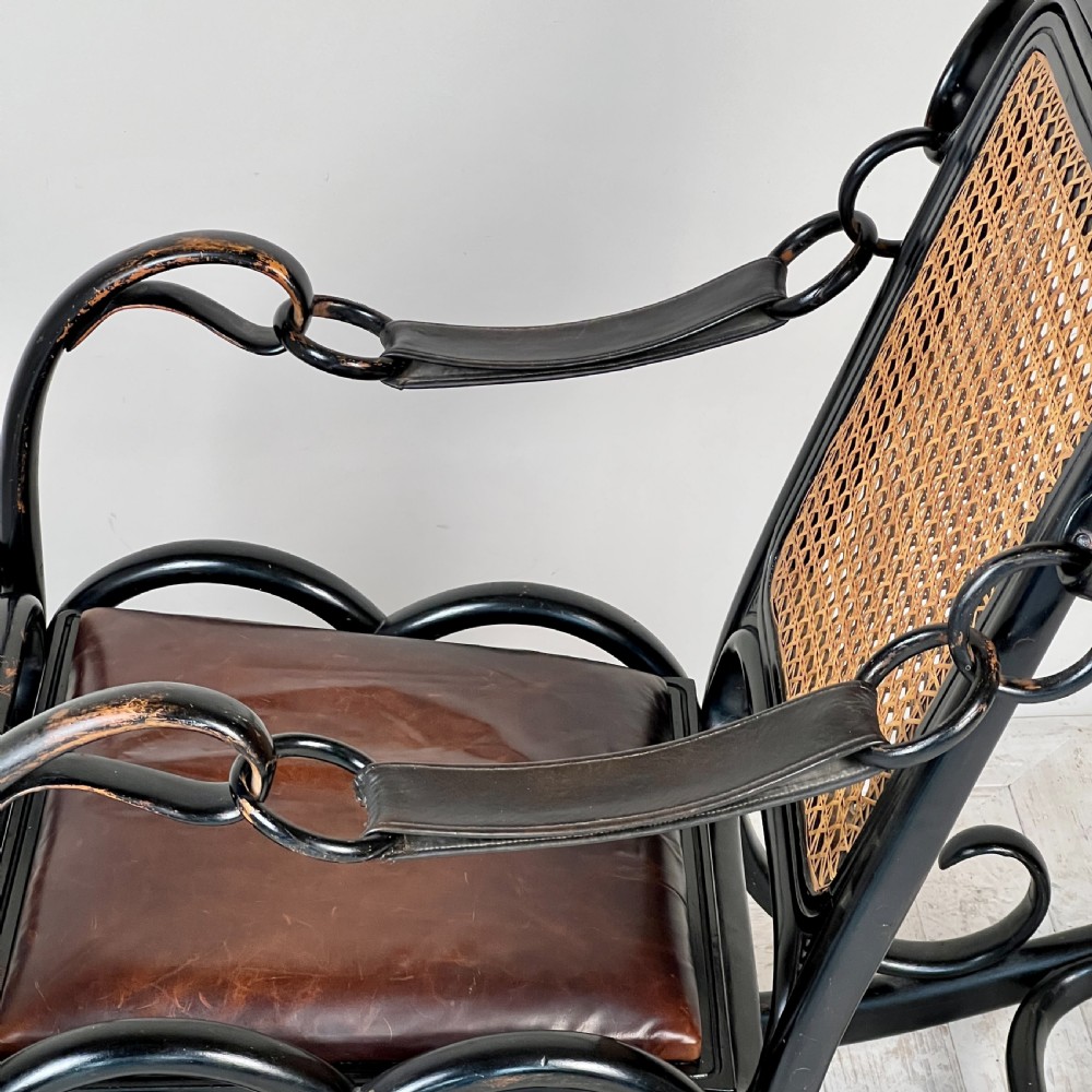 A dark brown steam-bent beechwood rocking chair featuring elaborate side scrollwork, a caned backrest, leather arm straps, and a dark leather padded seat. View VI
