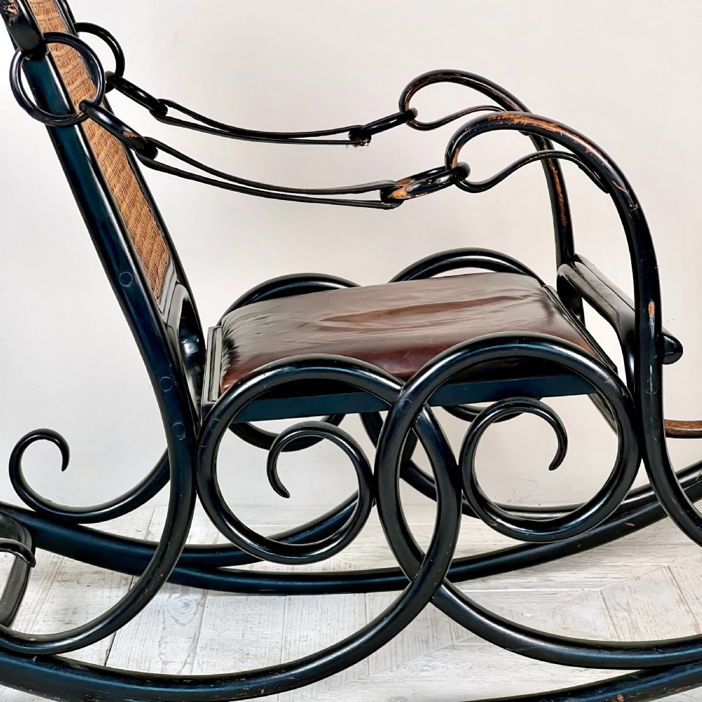 A dark brown steam-bent beechwood rocking chair featuring elaborate side scrollwork, a caned backrest, leather arm straps, and a dark leather padded seat. View VII