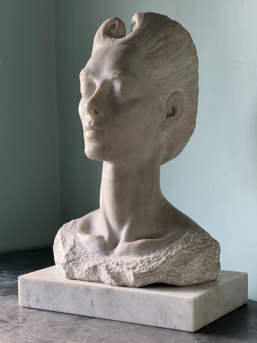 A carved off-white marble bust depicting a lady with styled hair and a serene expression, mounted on a matching square marble plinth. View VIII