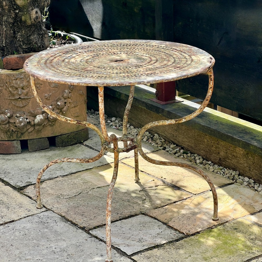 A circular French antique wrought iron garden table featuring a perforated metal top, curved legs with a central stretcher ring, and a heavily patinated weathered paint finish.
