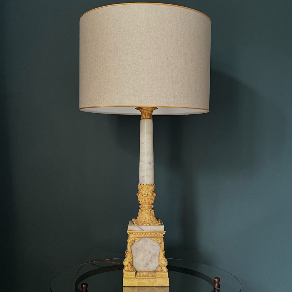 An Empire style table lamp featuring a white marble column base with gold ormolu leaf mounts and a large cream drum shade.