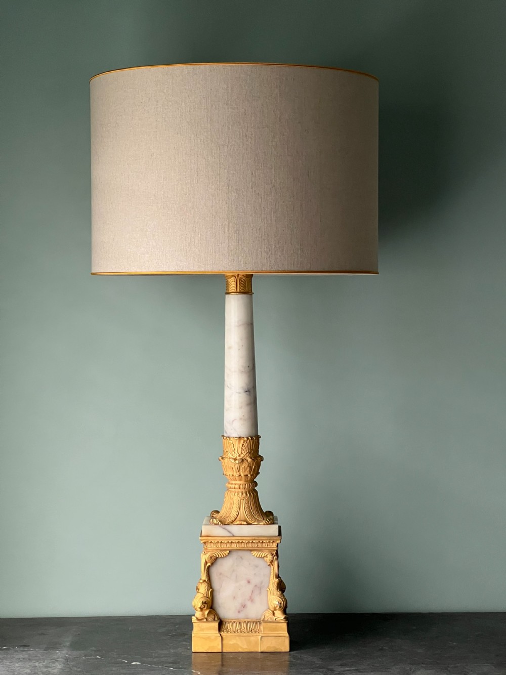 An Empire style table lamp featuring a white marble column base with gold ormolu leaf mounts and a large cream drum shade. View II