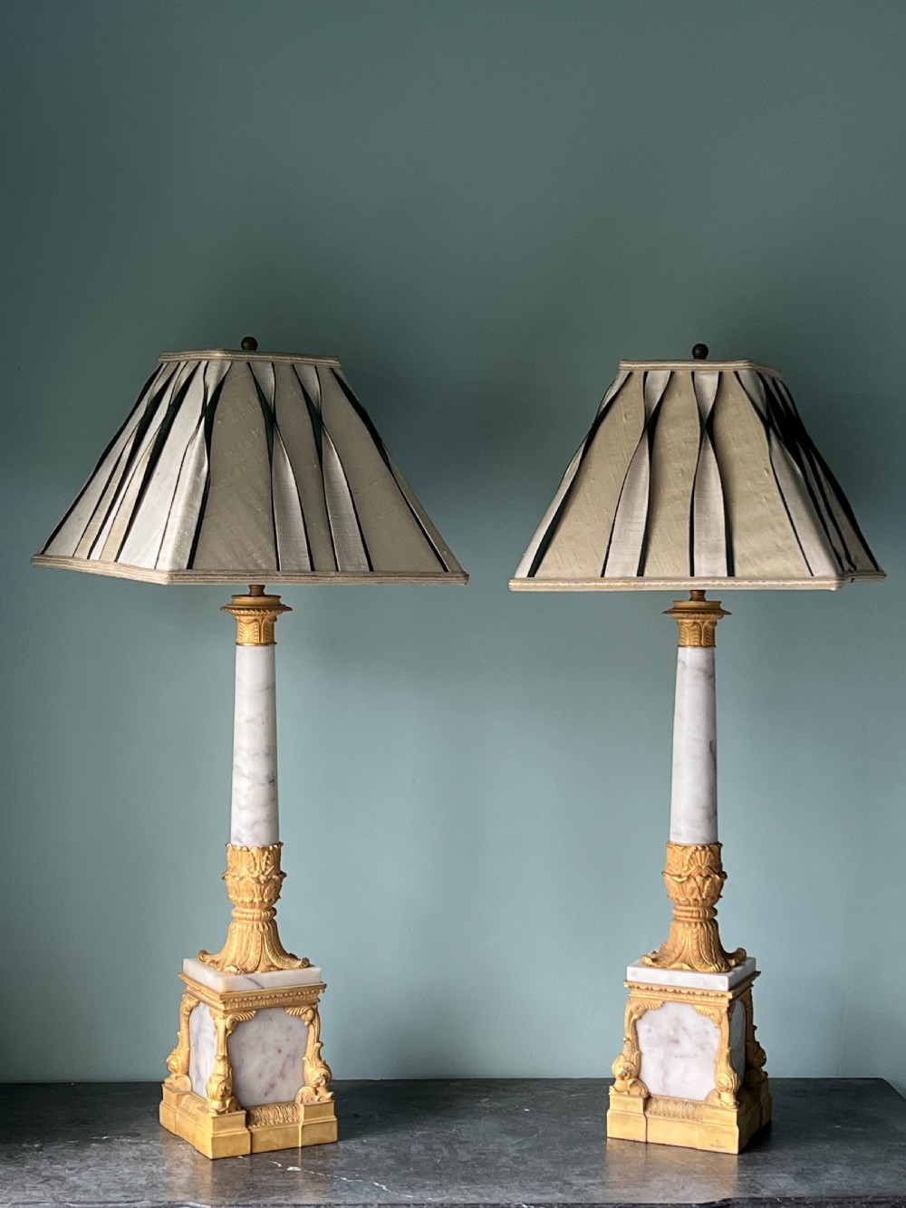 An Empire style table lamp featuring a white marble column base with gold ormolu leaf mounts and a large cream drum shade. View X