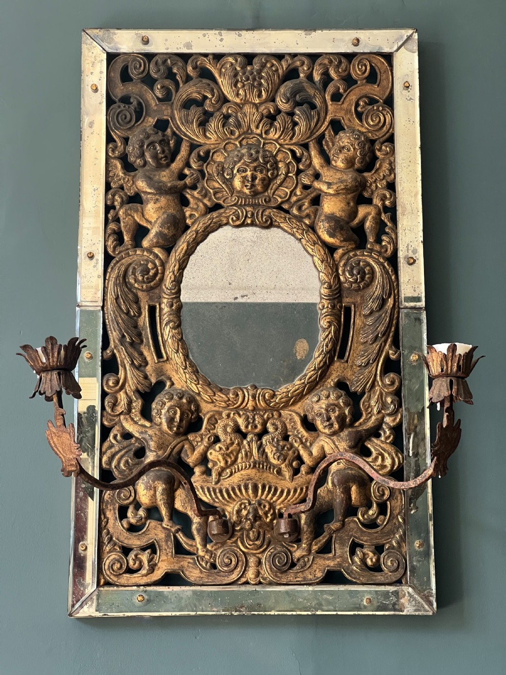 A rectangular gilded wall mirror featuring a central oval plate surrounded by relief carvings of putti and laurel leaves, flanked by two metal candle sconces.