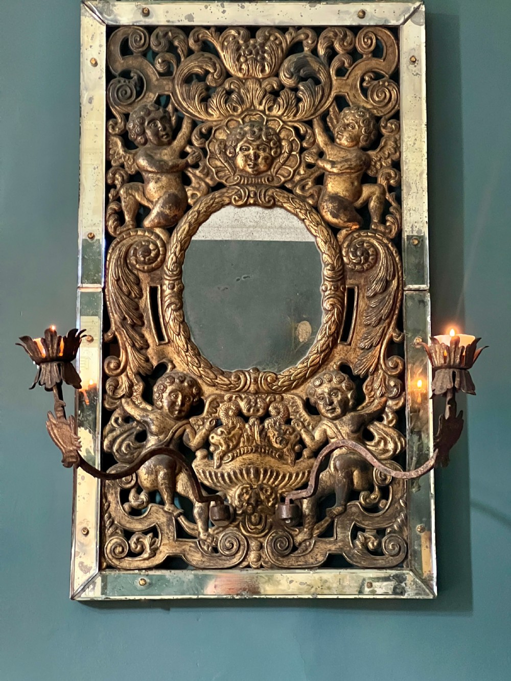 A rectangular gilded wall mirror featuring a central oval plate surrounded by relief carvings of putti and laurel leaves, flanked by two metal candle sconces. View II