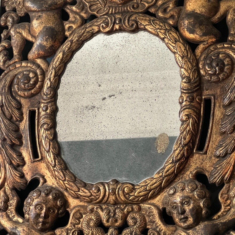 A rectangular gilded wall mirror featuring a central oval plate surrounded by relief carvings of putti and laurel leaves, flanked by two metal candle sconces. View III