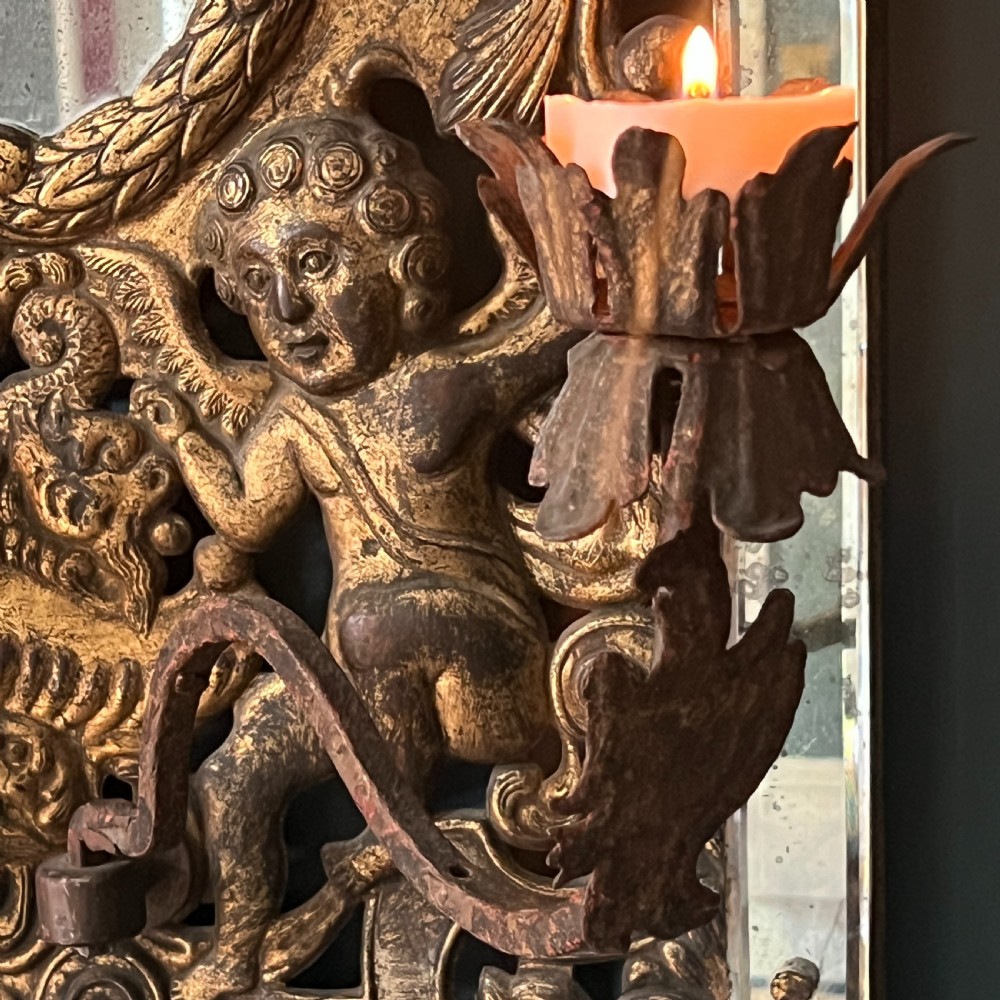 A rectangular gilded wall mirror featuring a central oval plate surrounded by relief carvings of putti and laurel leaves, flanked by two metal candle sconces. View IV