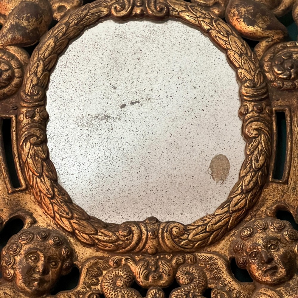 A rectangular gilded wall mirror featuring a central oval plate surrounded by relief carvings of putti and laurel leaves, flanked by two metal candle sconces. View V