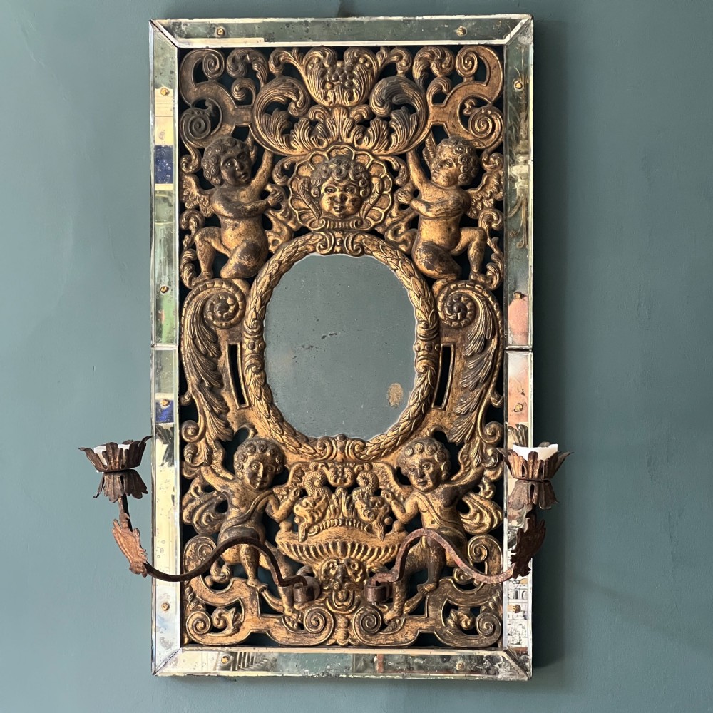 A rectangular gilded wall mirror featuring a central oval plate surrounded by relief carvings of putti and laurel leaves, flanked by two metal candle sconces. View VI
