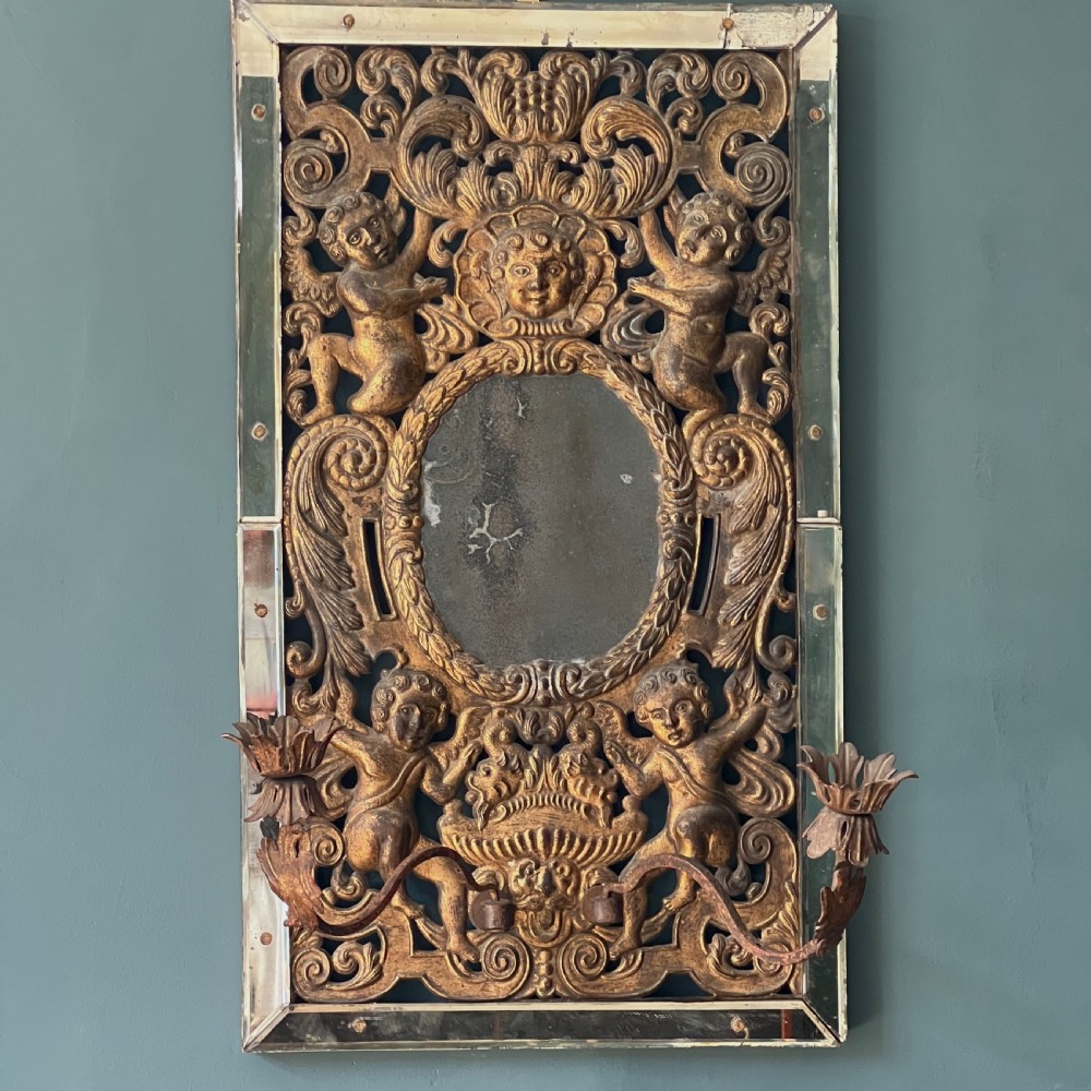 A rectangular gilded wall mirror featuring a central oval plate surrounded by relief carvings of putti and laurel leaves, flanked by two metal candle sconces. View VIII
