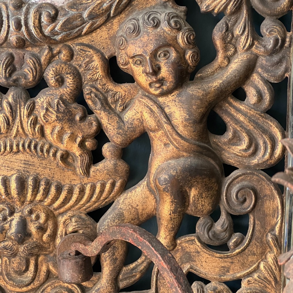 A rectangular gilded wall mirror featuring a central oval plate surrounded by relief carvings of putti and laurel leaves, flanked by two metal candle sconces. View IX