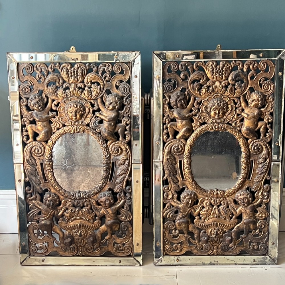 A rectangular gilded wall mirror featuring a central oval plate surrounded by relief carvings of putti and laurel leaves, flanked by two metal candle sconces. View X