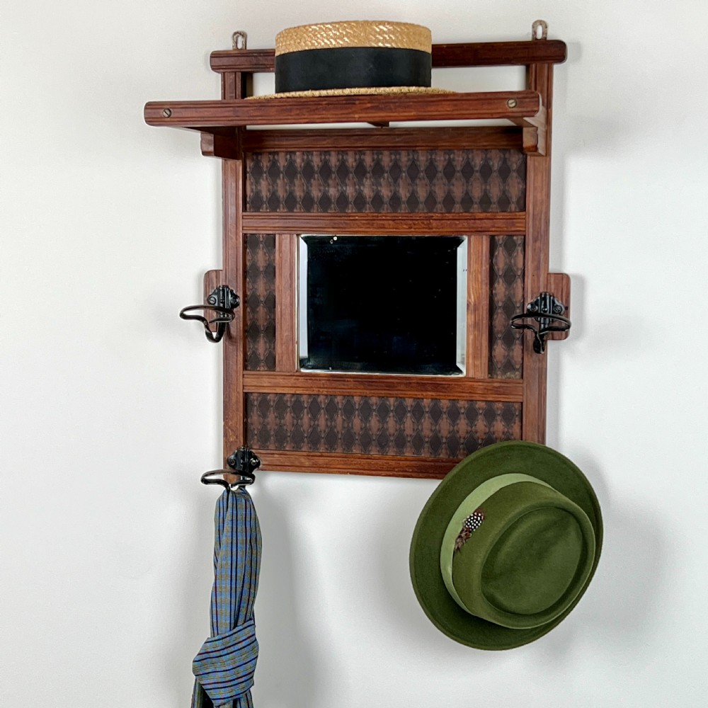 A wall-mounted dark wood coat rack featuring a central bevelled square mirror, geometric diamond pattern panels, an upper hat shelf, and four metal hooks. View III