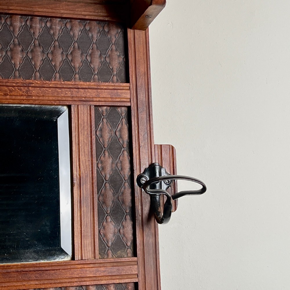 A wall-mounted dark wood coat rack featuring a central bevelled square mirror, geometric diamond pattern panels, an upper hat shelf, and four metal hooks. View V