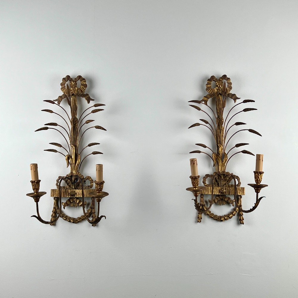 A pair of early 20th-century Italian giltwood and toleware wall sconces featuring carved ribbon bow crests, rising foliage sprigs, and twin candle arms.