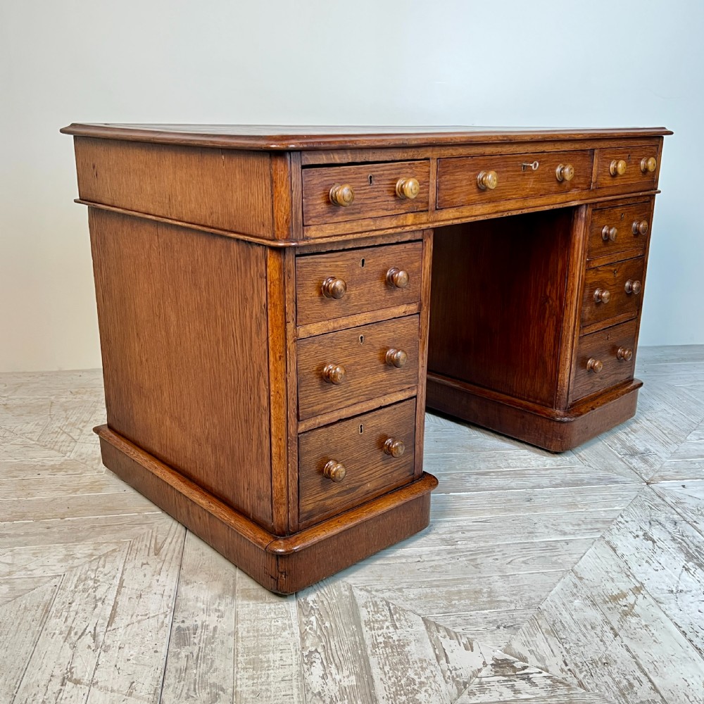 An antique Victorian oak pedestal desk featuring nine drawers with turned wooden knobs and a leather writing surface on twin plinth bases. View VI