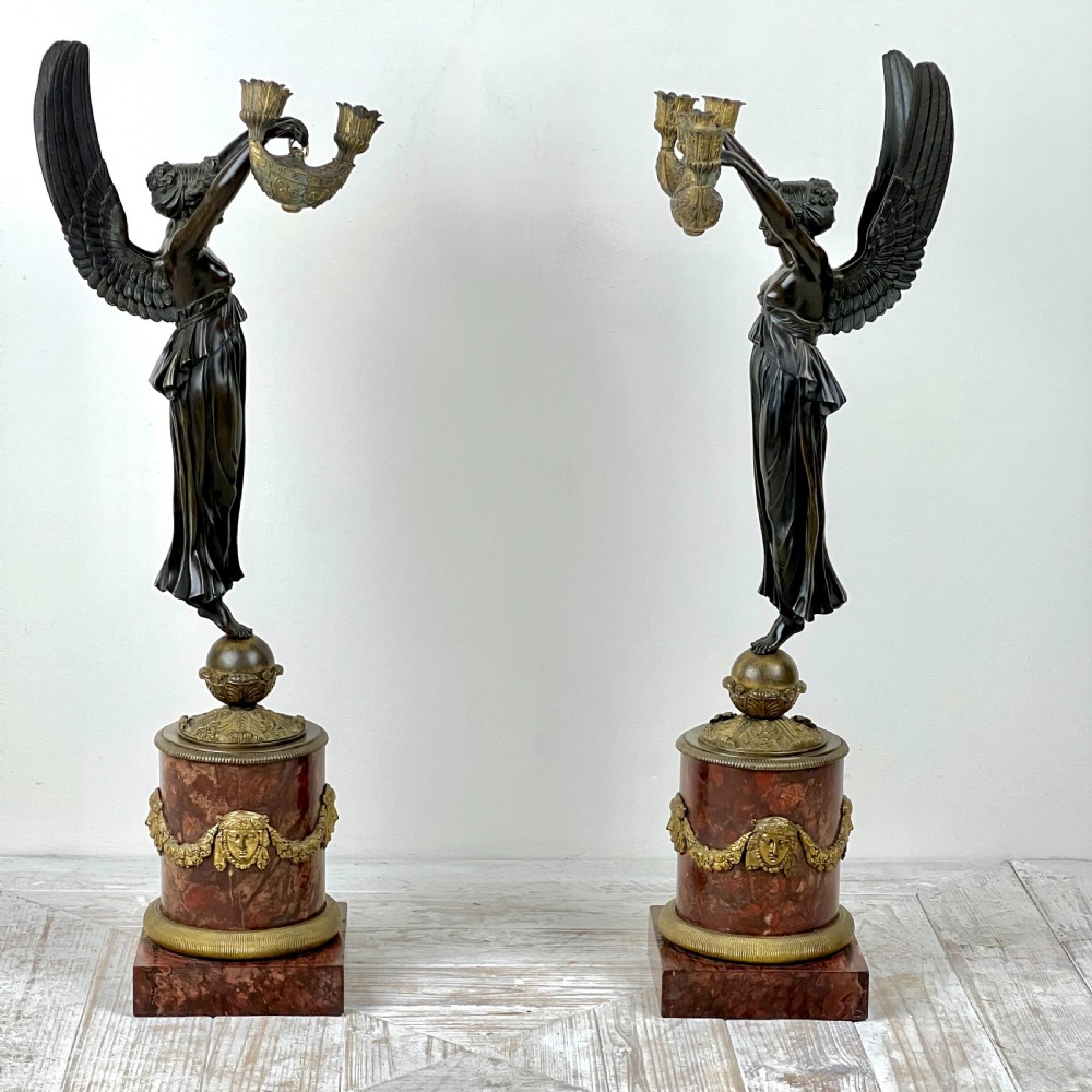 A pair of French Empire patinated bronze candelabra depicting winged victory figures standing on spheres atop rouge marble plinths with gilt bronze mounts.
