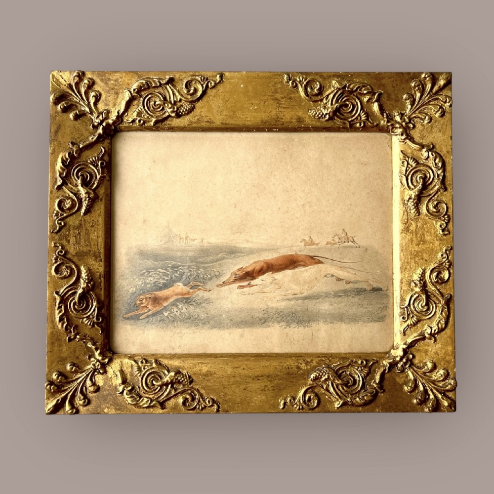 A 19th-century watercolour painting depicting two greyhounds pursuing a hare across a field, housed in a wide, ornate gilt frame with acanthus corner details.